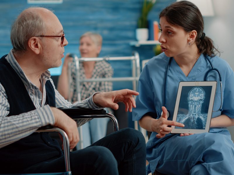Medical assistant holding tablet with x ray scan for checkup with disabled man in nursing home. Nurse explaining radiography on digital device to senior patient sitting in wheelchair